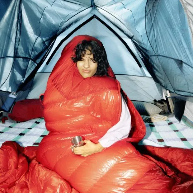 Jeune femme dans un sac de couchage rouge, assise avec une tasse sous la tente, plissant les yeux au soleil du matin