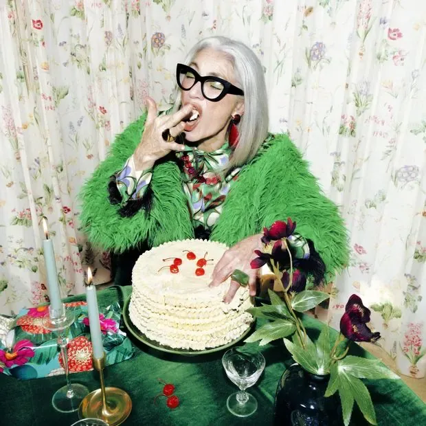Femme aux cheveux gris et à la veste à plumes verte mangeant un gâteau à sa table d’anniversaire